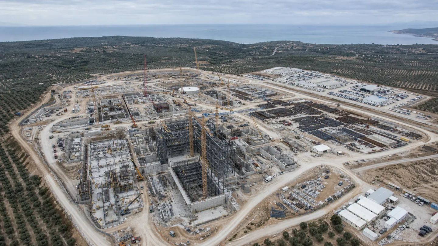 Αεροφωτογραφία μεγάλου εργοταξίου σταθμού: θεμελιώσεις, χώρος εναπόθεσης υλικών, γερανοί και εκατοντάδες οχήματα, μέσα σε ξηρό μεσογειακό τοπίο με ελαιώνες.